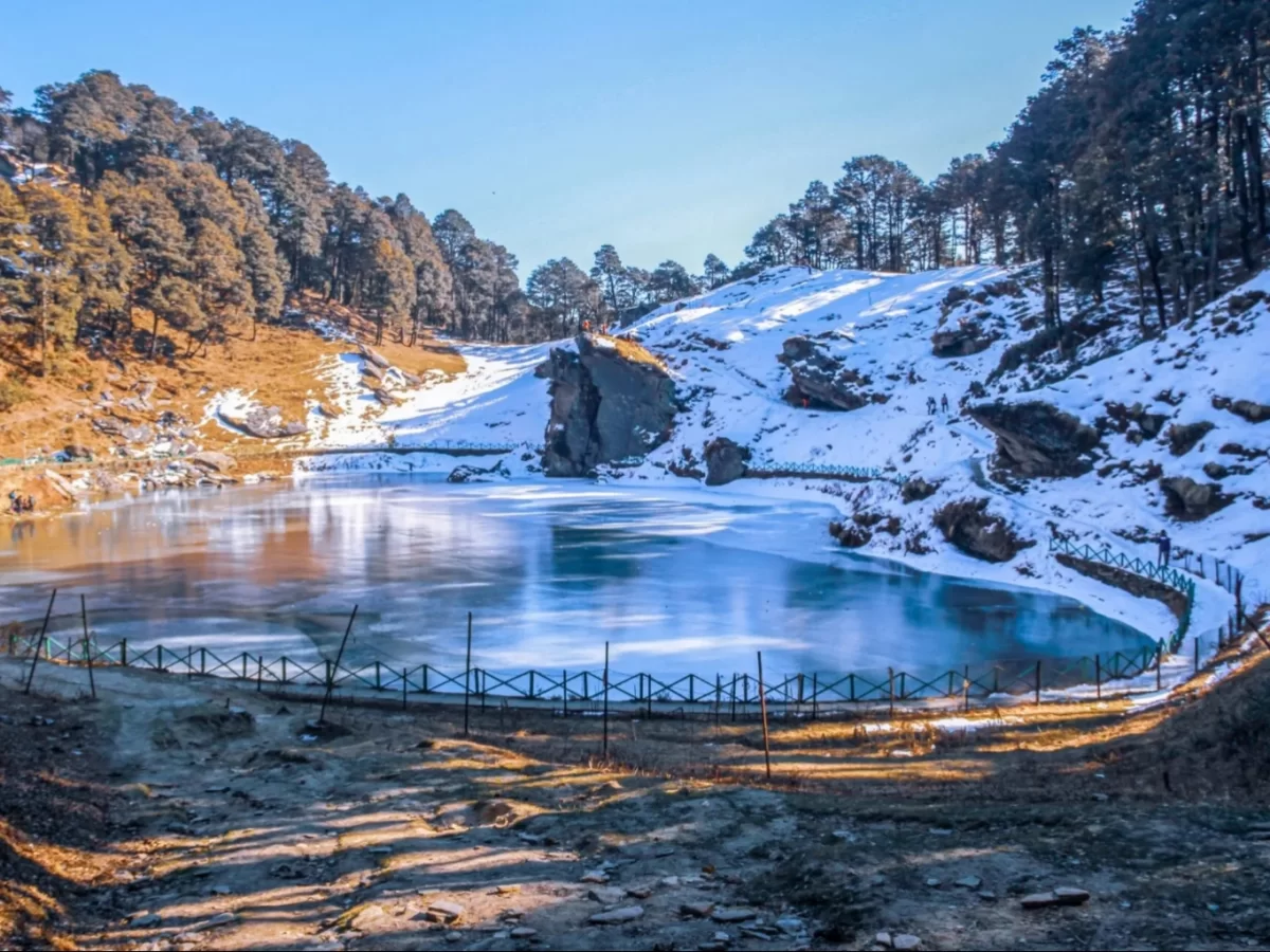 Frozen Serolsar Lake Jibhi during winter sunny day, featuring deodar forests, rocky cliffs, snow patches, railing path, perfect serene Himachal Pradesh tour package.
