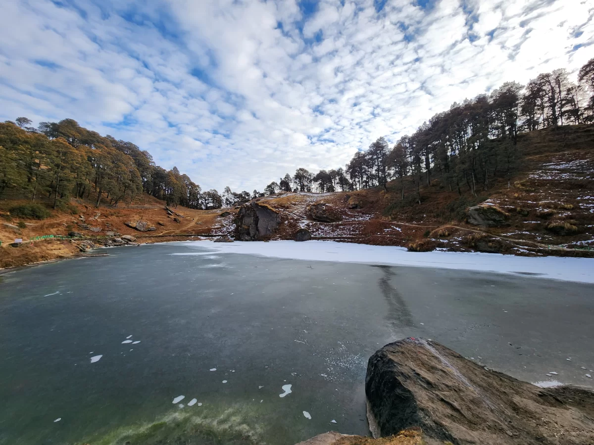 Frozen Serolsar Lake at Jibhi during winter partly cloudy, featuring icy surface, oak forests, rocky hills, perfect serene Himachal Pradesh tour package.