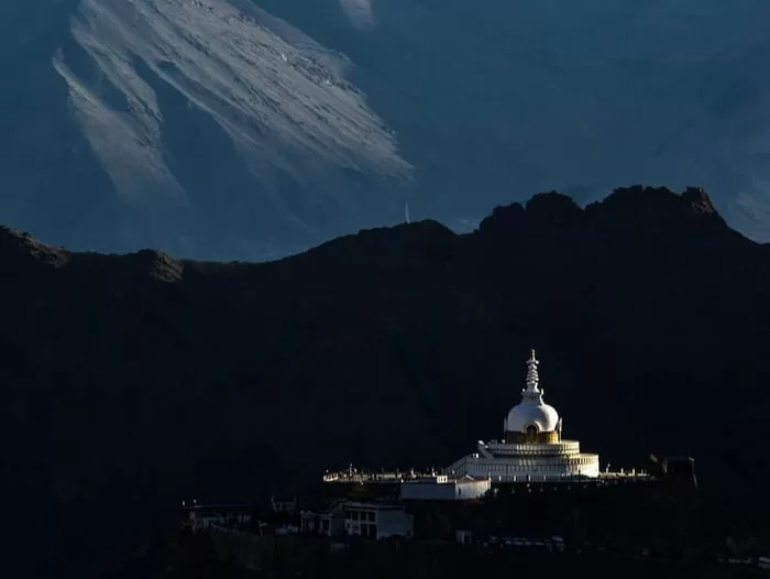 Shanti Stupa Leh Ladakh at twilight with snowy peak backdrop, featuring illuminated white stupa with golden spire, dramatic clouds, rugged mountains, perfect spiritual sunset panoramic experience with Leh Ladakh sightseeing tour package.
