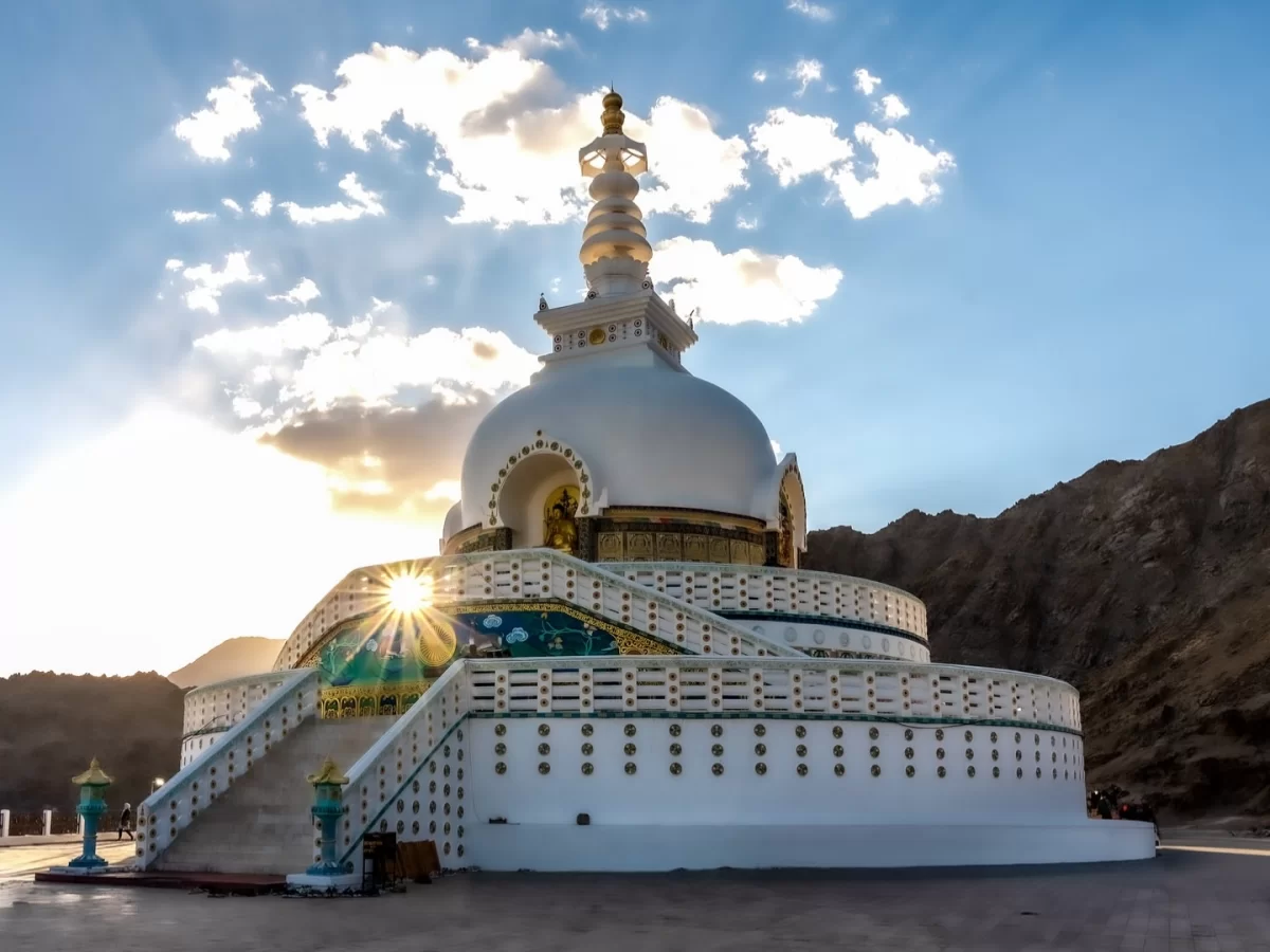Shanti Stupa Leh Ladakh bathed in golden sunset sunlight amid partly cloudy sky, featuring white dome stupa golden spire, colorful base, rugged mountain backdrop, perfect serene spiritual sunset experience with Leh Ladakh sightseeing tour package.