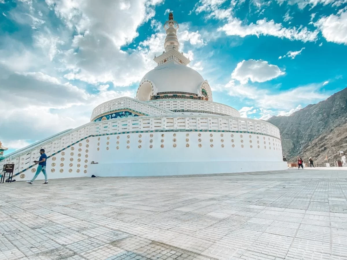 Wide angle Shanti Stupa Leh Ladakh under partly cloudy blue sky, featuring white stupa dome spire, colorful base railings, tourists walking stairs plaza, rugged mountain backdrop, perfect panoramic spiritual experience with Leh Ladakh sightseeing tour pac
