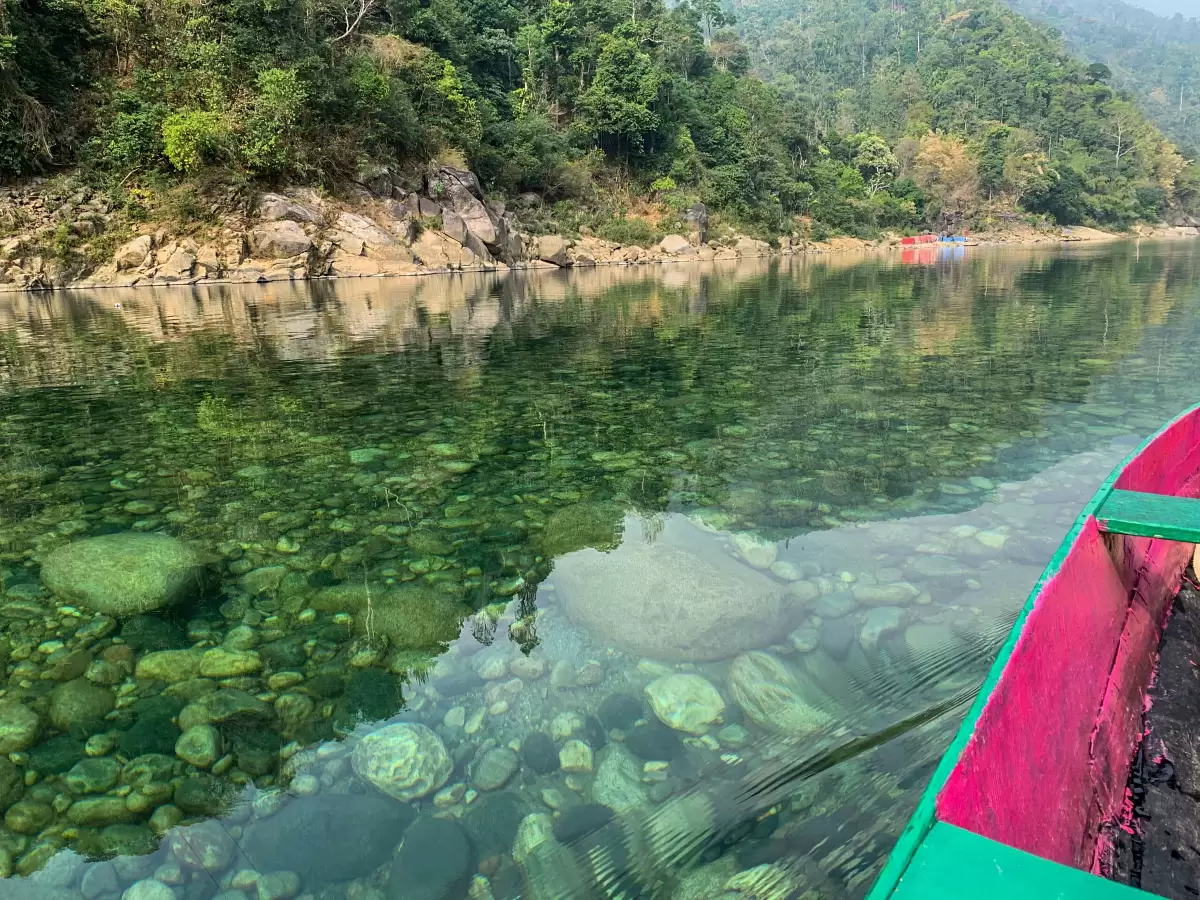 Shnongpdeng Village near Dawki Meghalaya Umngot River pink green wooden boat view crystal clear emerald green waters visible riverbed pebbles rocks lush green forested hills serene tranquil adventure perfect boating camping rafting Meghalaya tour package.