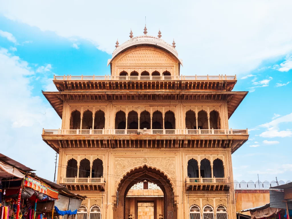 Ornate haveli-style gate of Sri Rangji Mandir during partly cloudy daytime, featuring multi-tiered sandstone architecture, bustling market surroundings, perfect spiritual heritage experience with Vrindavan tour package