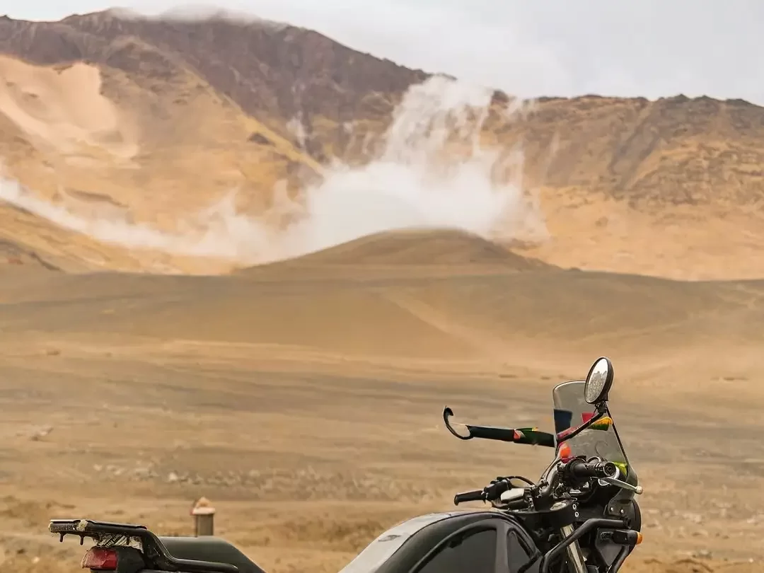 Royal Enfield motorcycle parked near Siachen Base Camp in Ladakh during dusty sunset, featuring sand dunes, hazy mountains, perfect adventure bike Siachen tour package. 