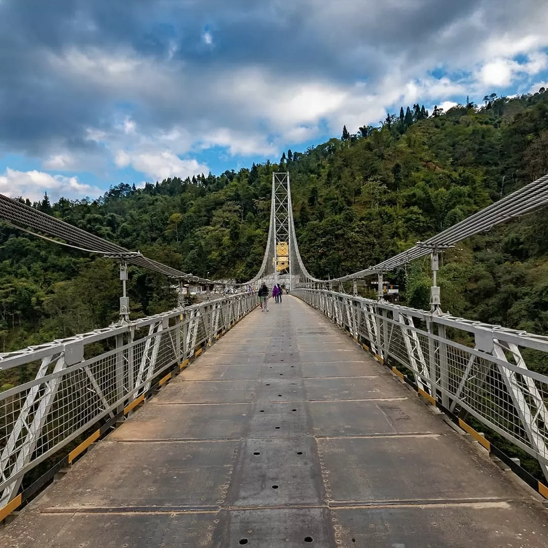 Couple walking on Singshore Bridge Pelling under dramatic cloudy sky, surrounded by lush green hills, perfect adventure Sikkim tour package.