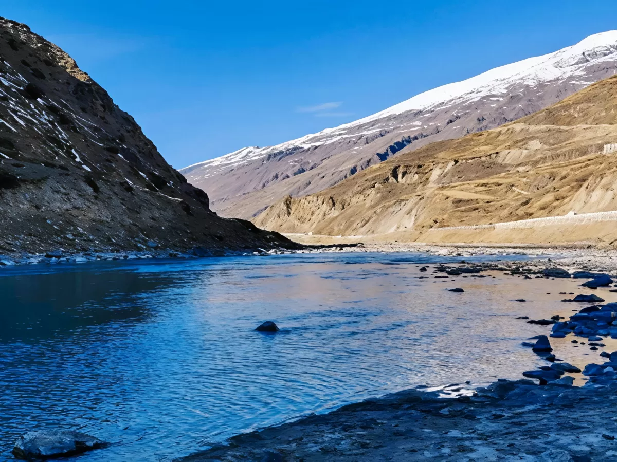 Sissu Valley serene turquoise river meandering through barren brown canyon walls with light snow patches on distant peaks under clear blue skies, rocky shores, perfect Himachal Pradesh adventure tour package.