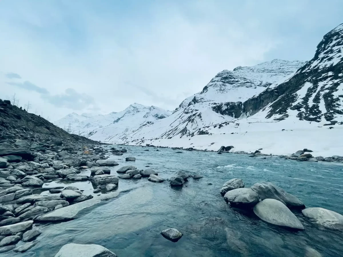 Sissu Valley turquoise glacial river rushing over massive boulders amid snow-blanketed towering peaks under cloudy blue skies, rocky banks, perfect Himachal Pradesh adventure tour package.