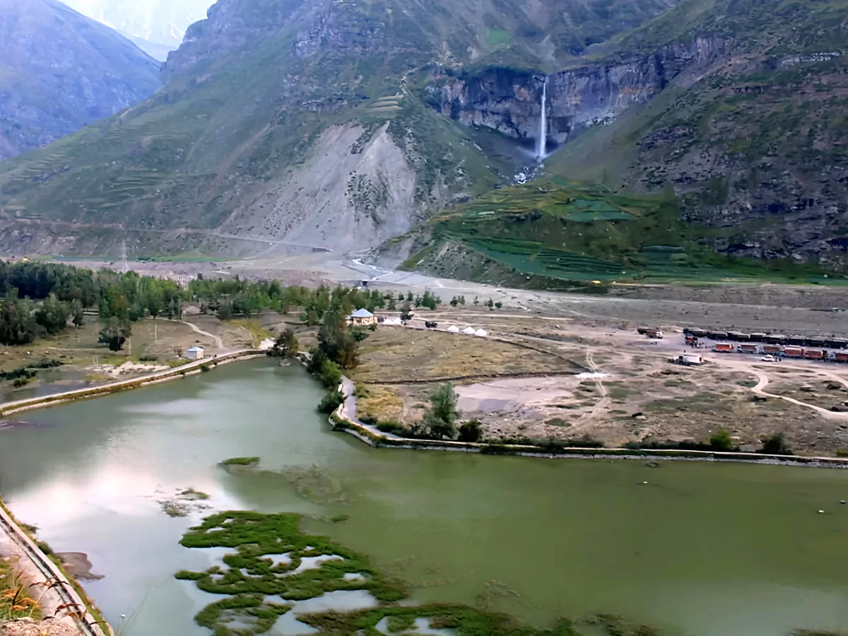 Sissu Lake lush green valley with prominent waterfall cascading from cliff amid terraced fields, green poplar groves, tent structures, trains, algae-covered waters bordered by paths, perfect Himachal Pradesh tour package.