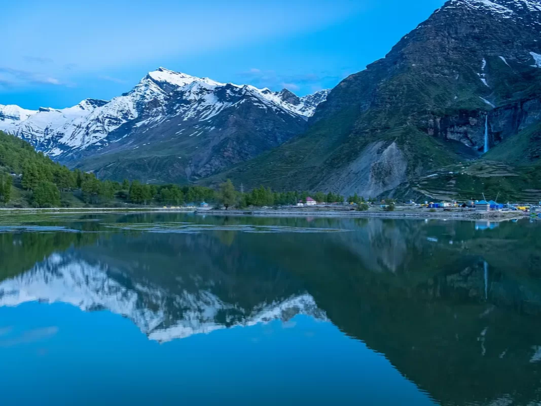 Sissu Lake mirror reflection of snow-capped peaks, cascading waterfall, colorful village huts with green trees along deep blue waters under twilight blue sky, perfect Himachal Pradesh tour package.