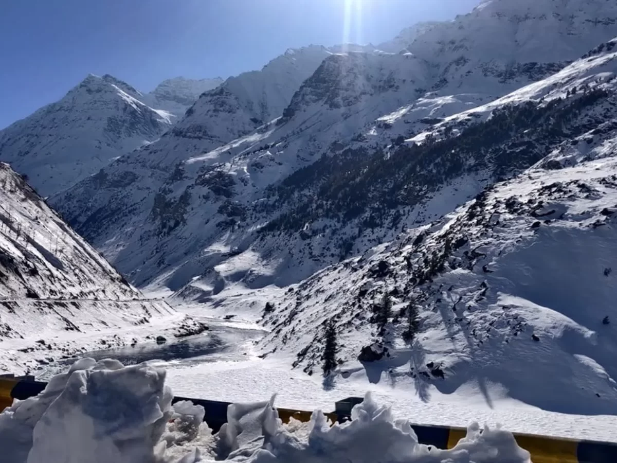 Sissu Lake en route with foreground yellow-blue prayer flags and piled snow against radiant sunlit snow peaks and valleys under deep blue sky, pine-dotted ridges, perfect Himachal Pradesh tour package.