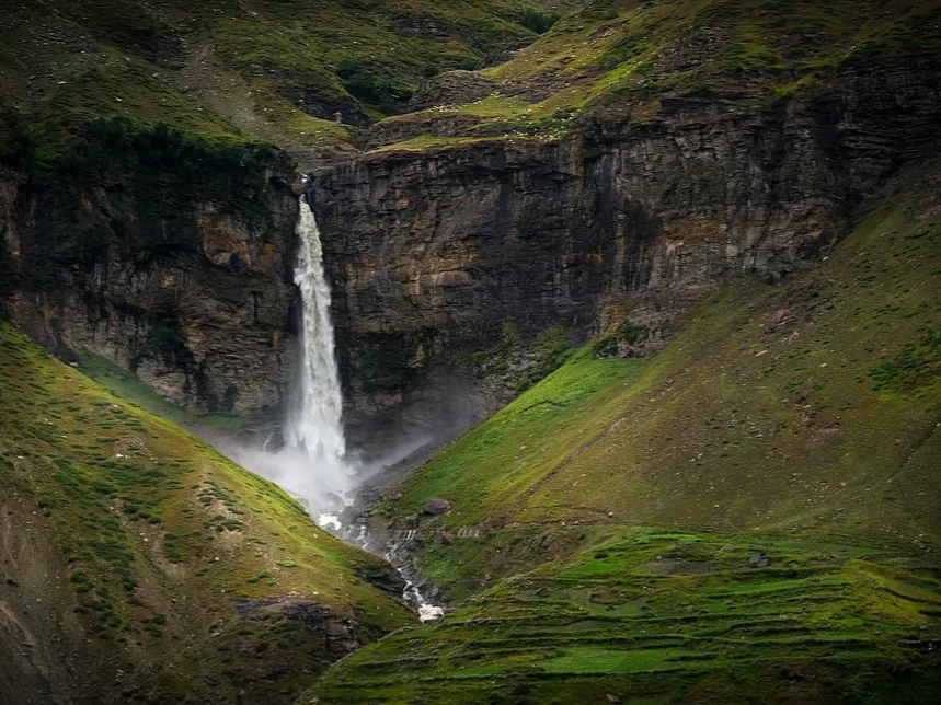 Sissu Waterfall cascading down a tall rocky cliff into a narrow stream below, surrounded by lush green hills and rugged mountain slopes in the Lahaul Valley.