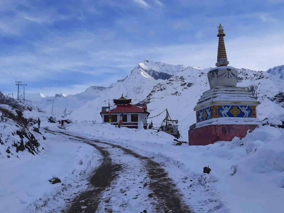 Snow-covered Sissu Monastery featuring a small temple with a red roof, a decorated Buddhist stupa in the foreground, winding path through deep snow, and towering Himalayan peaks in the background.