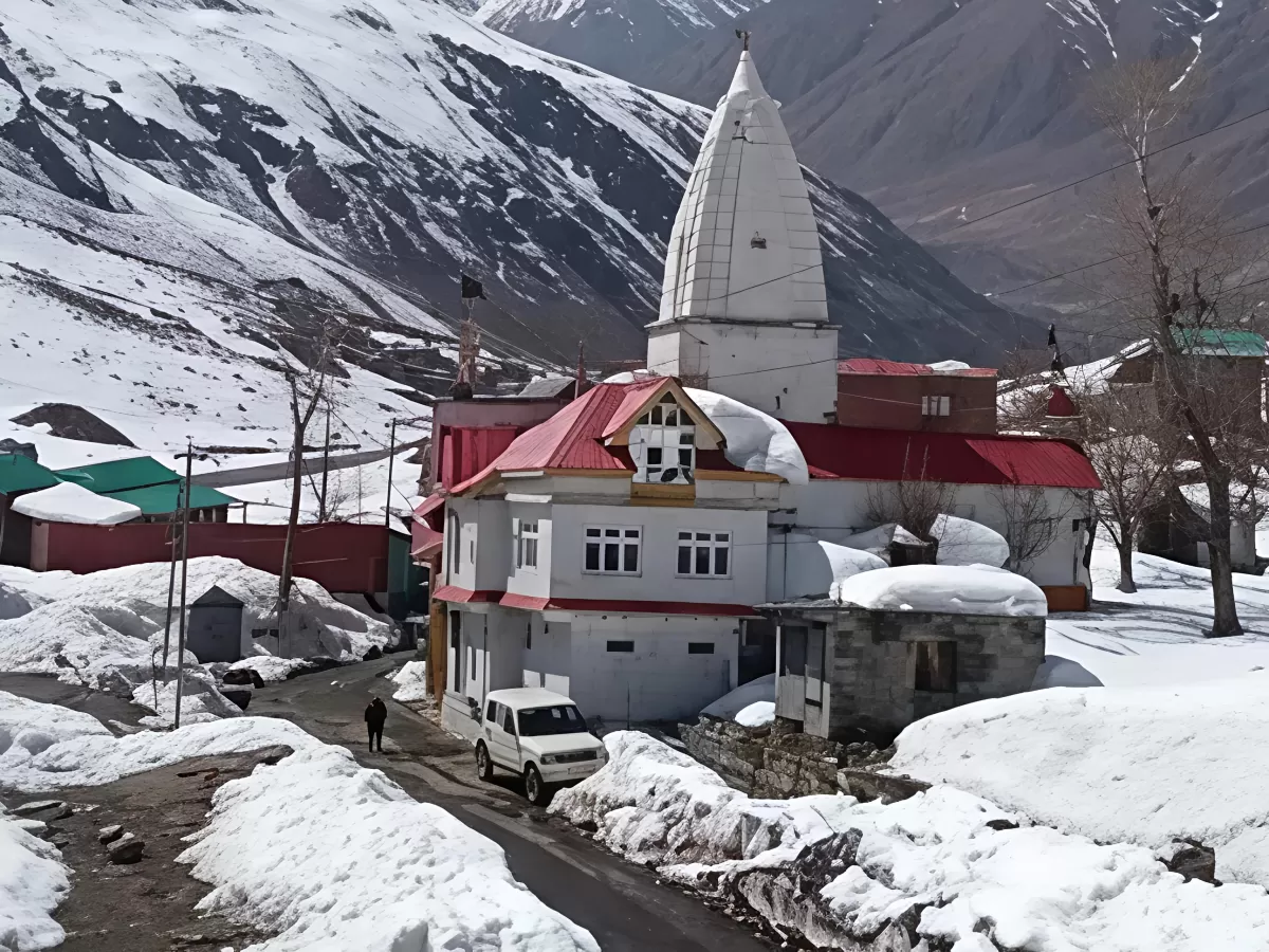 Sissu Raja Ghepan Temple snowy Lahaul village red roof white shikhara temple complex parked white SUV local walking snow-covered road majestic snow-capped peaks backdrop, perfect Himachal tour packages.