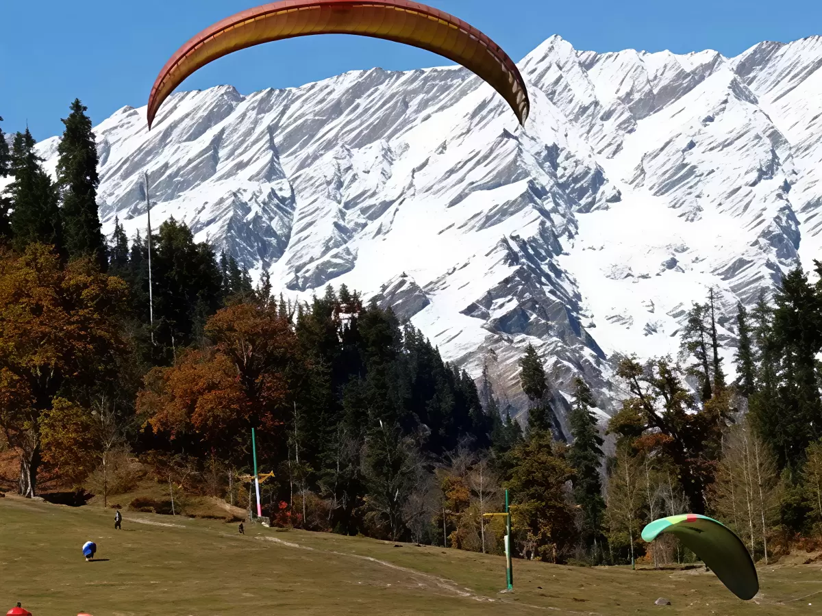 Paragliding adventure at Solang Valley Manali during sunny skies, featuring orange green gliders over snowy peaks pine forests and meadows, perfect adventure experience Himachal Pradesh tour package.