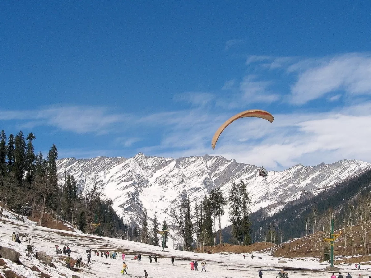 Paragliding at Solang Valley Manali during winter clear sky, featuring orange glider, snowy slopes, pine trees, crowds, perfect adventure Himachal Pradesh tour package.