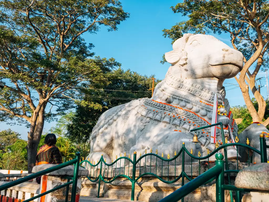 Sri Nandi Temple in Mysore during sunny daytime, featuring massive white Nandi statue beneath green trees, perfect spiritual Karnataka tour package
