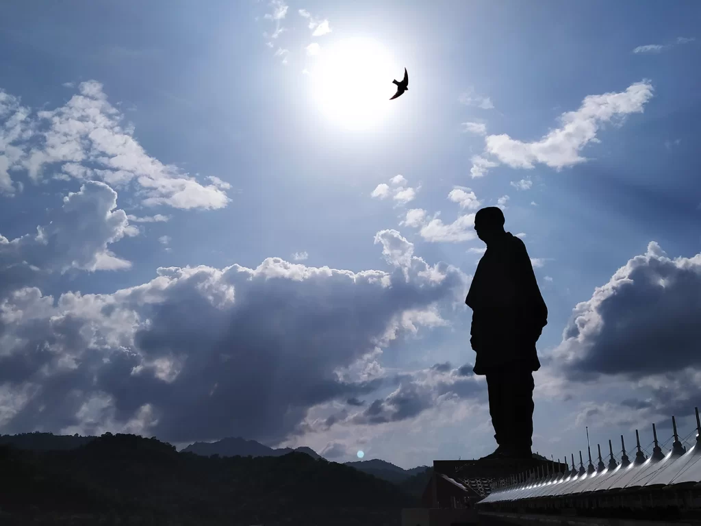 Statue of Unity silhouette during golden hour in Gujarat, featuring backlit Sardar Patel figure, flying bird, radiant sun, white canopies and surrounding hills, perfect cultural landmark with Gujarat tour packages.