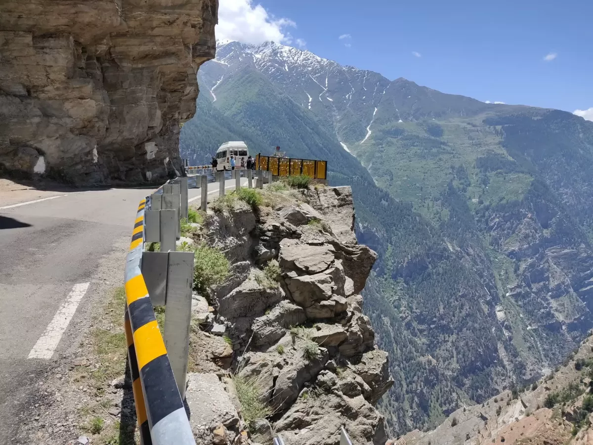 Suicide Point Kalpa Kinnaur during clear skies, featuring roadside viewpoint vehicles barriers dramatic cliff drop lush green valley snow mountains, perfect adventure viewpoint Himachal Pradesh tour package.