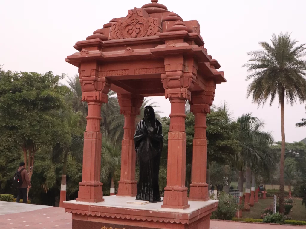 Sun Temple Gwalior temple complex during clear afternoon, featuring red sandstone pavilion with black statue of Smt. Mahadevi Birla in prayer pose amid palm trees and gardens, perfect heritage experience in Madhya Pradesh tour package.