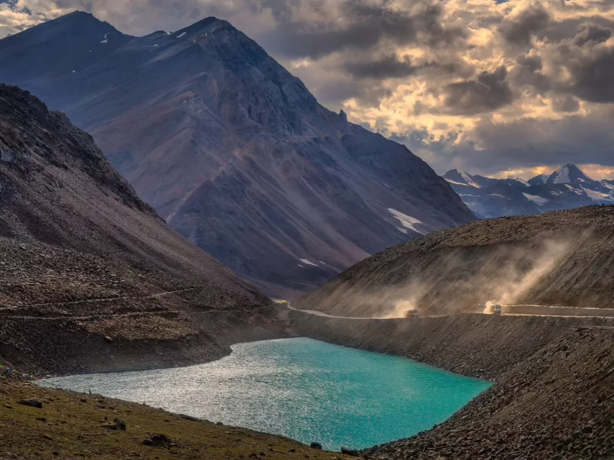 Dramatic Suraj Tal high-altitude lake below Baralacha La Pass on Manali Leh Highway Himachal Pradesh during golden hour sunset, featuring turquoise waters rugged mountains dust trail truck dramatic clouds snowy peaks, perfect road trip adventure experienc