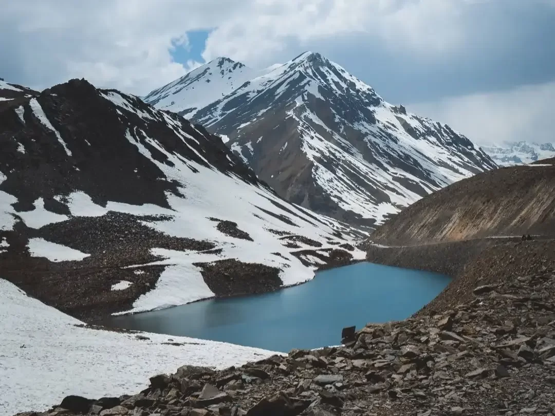 Suraj Tal Lahaul Spiti Himachal Pradesh India stunning blue glacial lake surrounded jagged snow capped peaks dark rocky slopes scattered snow patches cloudy skies foreground rocks highway, perfect Manali Leh highway breathtaking tour package.