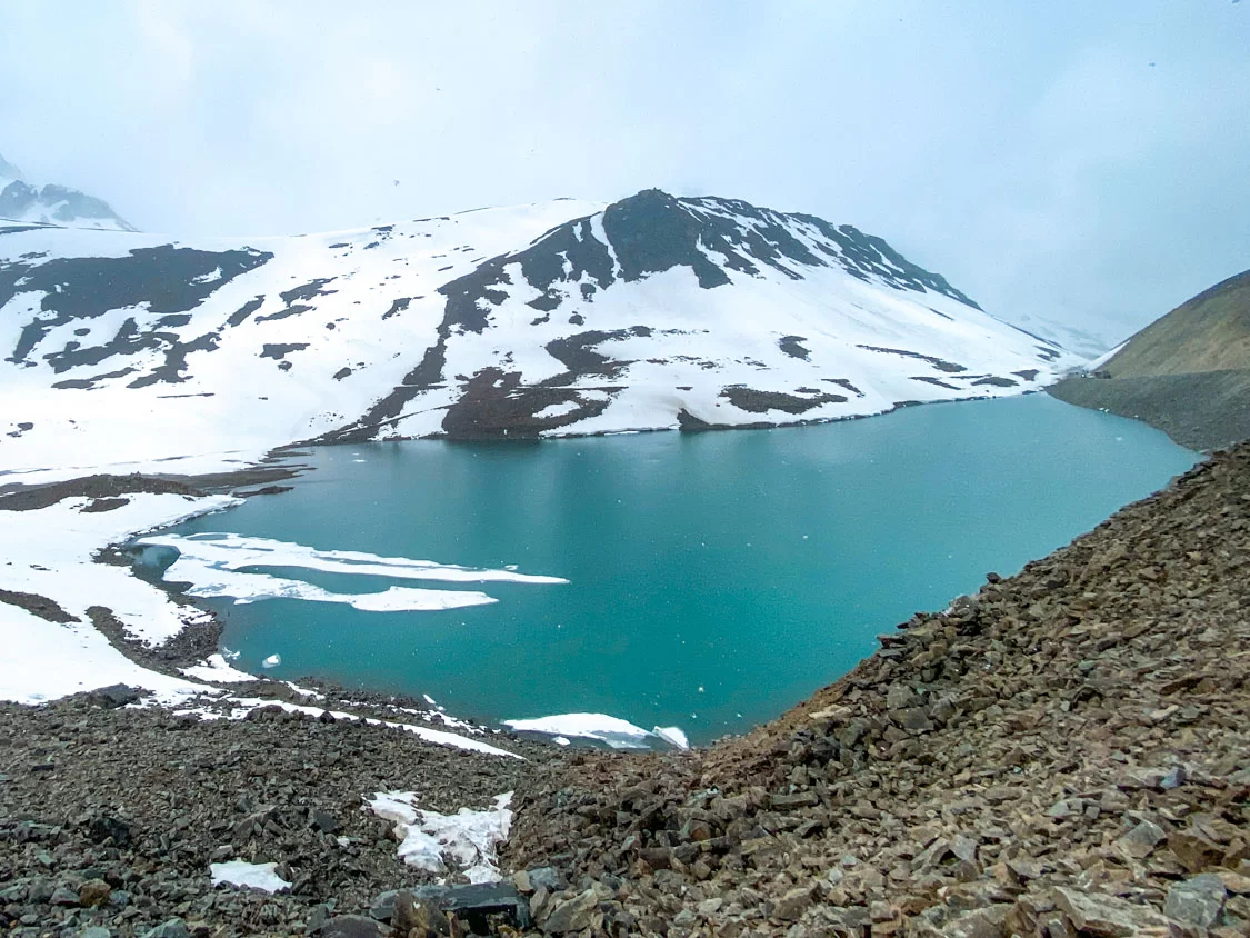 Suraj Tal Lahaul Spiti Himachal Pradesh India mesmerizing turquoise lake partially frozen amid heavy snow covered rugged mountains misty grey skies foreground rocky snow patches, perfect Manali Leh highway winter adventure tour package.
