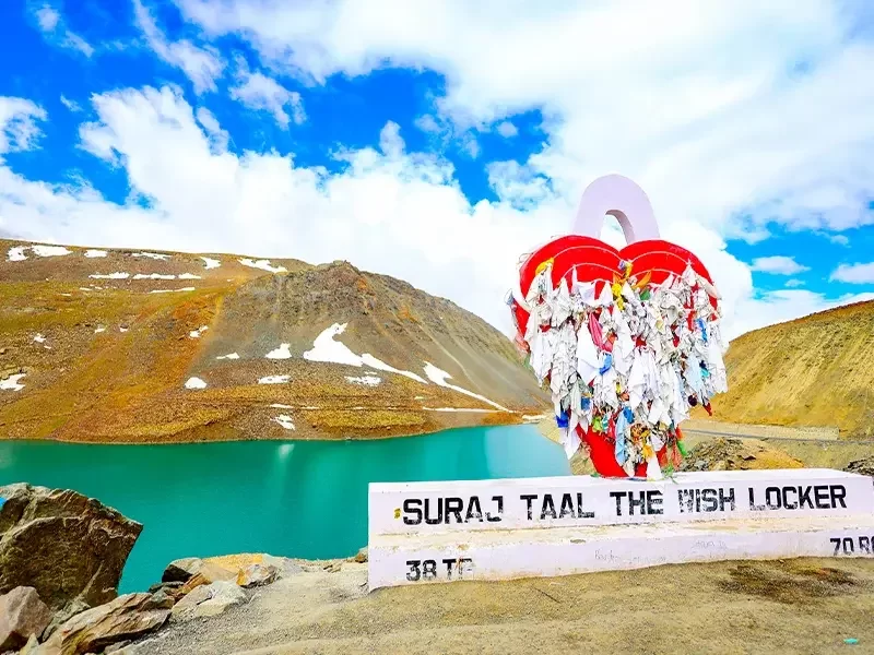 Suraj Tal Lahaul Spiti Himachal Pradesh India iconic red heart shaped wish locker signpost with colorful ribbons locks overlooking turquoise lake snowy brown mountains blue skies scattered clouds highway edge, perfect Manali Leh highway romantic tour pack