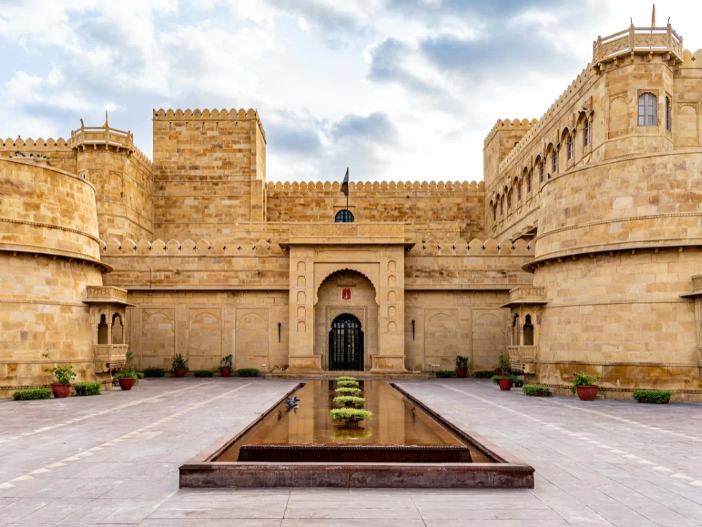 Fortress facade at Suryagarh Palace Jaisalmer during partly cloudy day, featuring central gate, flag, pool reflections and potted plants, perfect romantic experience in Rajasthan tour package. 