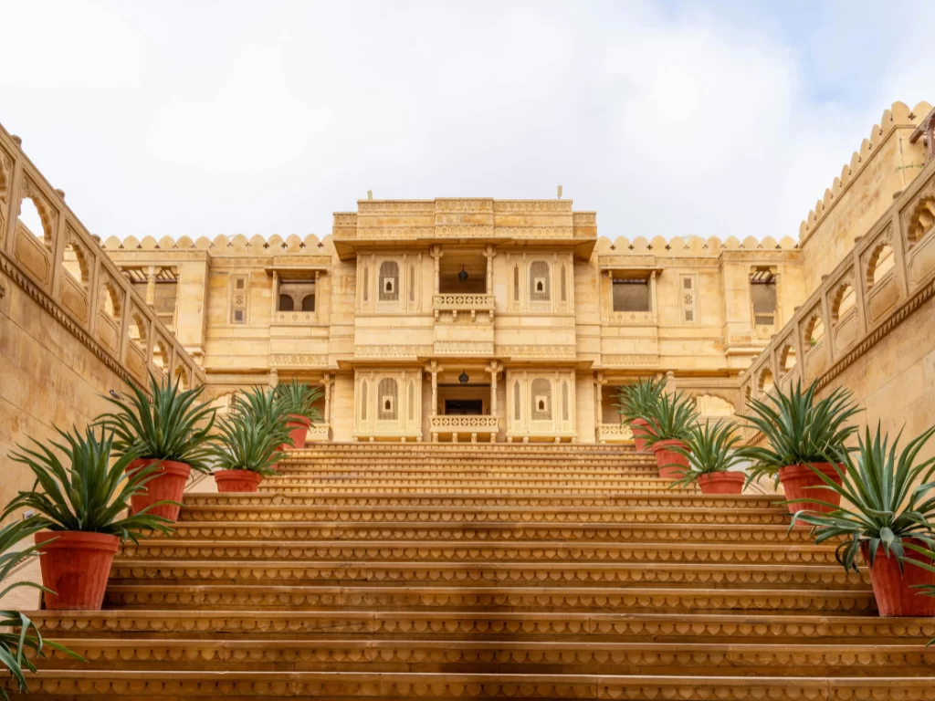 Grand entrance at Suryagarh Palace Jaisalmer during sunny day, featuring sandstone arches, steps and potted plants, perfect romantic experience in Rajasthan tour package.