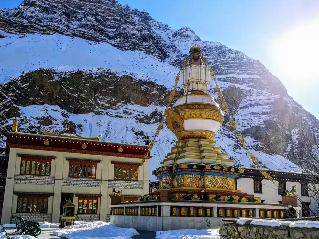 Golden prayer wheel at Tabo Monastery Spiti during winter sunny day, featuring snow peaks, Tibetan building, prayer flags, perfect spiritual Himachal Pradesh tour package.