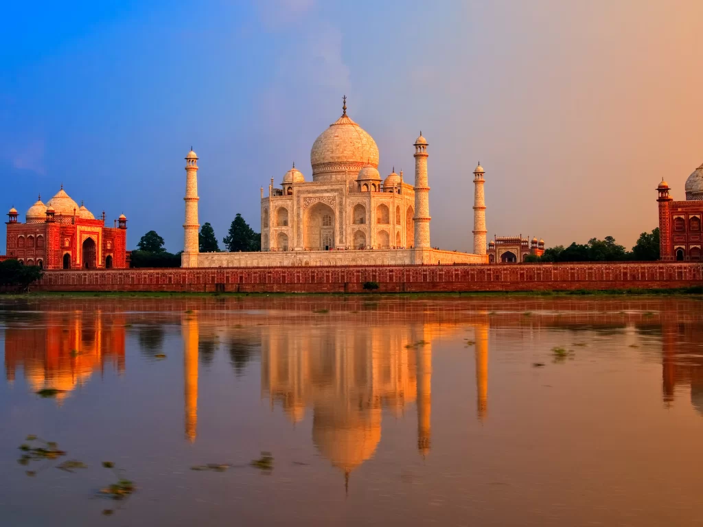 Taj Mahal Agra Uttar Pradesh perfect Yamuna River reflection at sunset with three domes mirrored in golden water, flanked by red sandstone buildings, blue-orange skies, perfect UNESCO heritage India tour package.