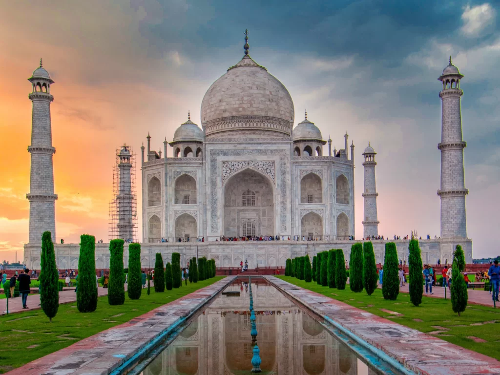 Taj Mahal Agra Uttar Pradesh during dramatic sunset with tourists, reflecting pool, cypress trees, white marble mausoleum, four minarets, orange-pink skies, perfect UNESCO heritage India tour package.