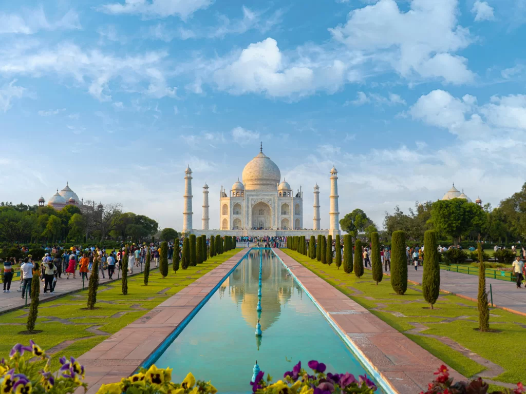 Taj Mahal Agra Uttar Pradesh wide angle reflection view with tourists, cypress avenue, perfect pool mirror image, white marble mausoleum, four minarets, blue skies with scattered clouds, perfect UNESCO heritage India tour package.