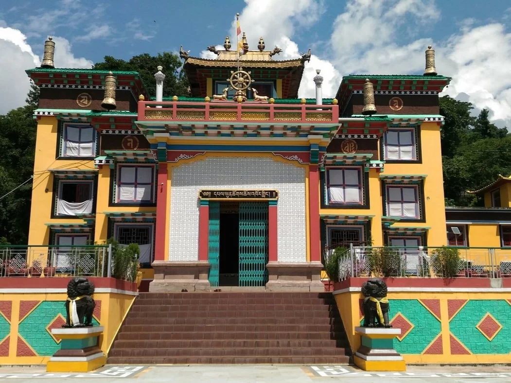 Front view of Tashi Thongyang Monastery featuring a vibrant yellow and red Tibetan-style facade, central entrance with decorative doorway, prayer symbols, rooftop ornaments, and lion statues at the staircase under a blue sky.