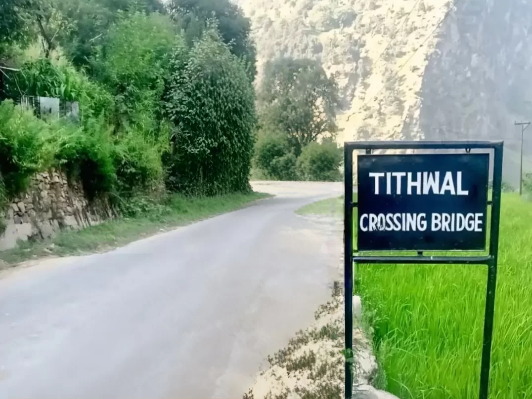 Teetwal Village Tithwal Crossing Bridge sign during sunny day Kashmir, featuring black frame road greenery rocky cliffs backdrop, perfect Kashmir tour packages.