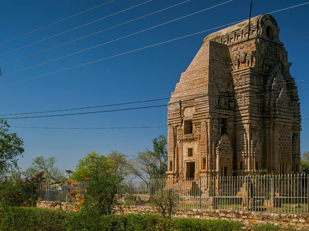 Teli ka Mandir Gwalior during clear afternoon, featuring side profile of towering phamsana shikhara with intricate sandstone carvings amid sparse vegetation and wire fencing, perfect heritage experience in Madhya Pradesh tour package.