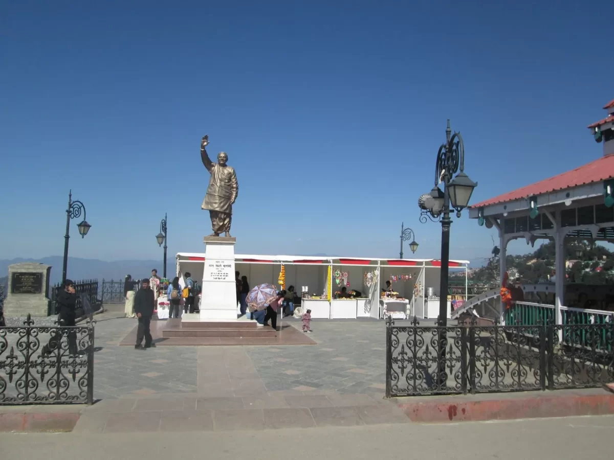 Gandhi Statue on The Ridge Shimla during sunny day, featuring bronze leader raising lamp pedestal surrounded tourists shops colonial lamp posts ridge view mountains background, perfect colonial heritage cultural experience Himachal Pradesh tour packages.