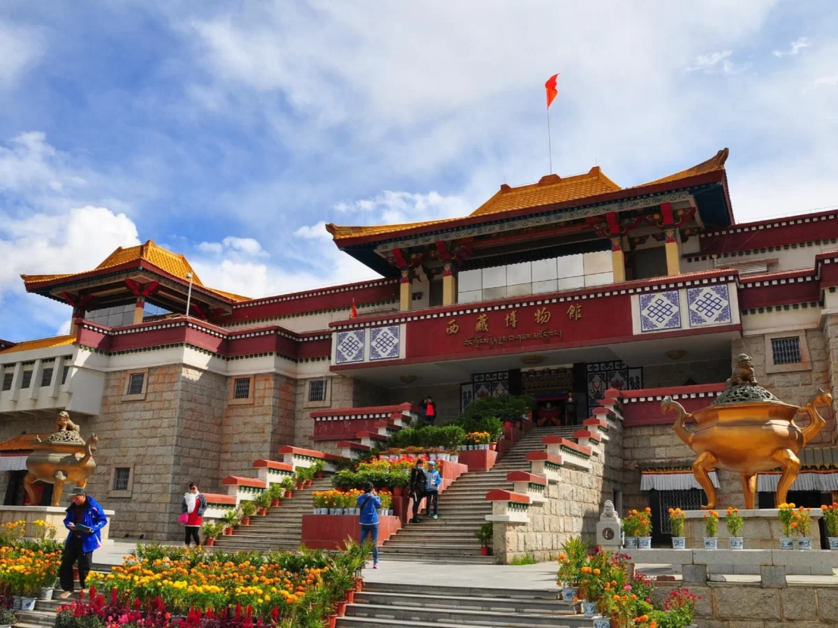 The Tibet Museum in Dharamshala featuring a grand Tibetan-style building with golden rooftops, red facade, wide staircase lined with flowers, and visitors at the entrance under a blue sky.