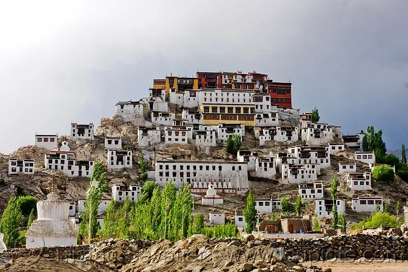 Thiksey Gompa multi-tiered monastery resembling Potala Palace near Leh Ladakh during cloudy day, featuring white buildings red golden roofs chortens green trees rocky terrain, perfect spiritual heritage experience with Thiksey Gompa Ladakh tour package. 