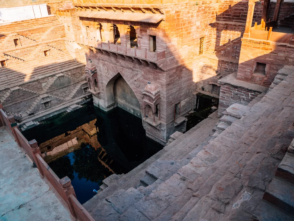Toorji Ka Jhalra Bavdi stepwell at Jodhpur during golden hour, featuring arched gateway, sandstone steps, water reflections, perfect heritage experience Rajasthan tour packages. 