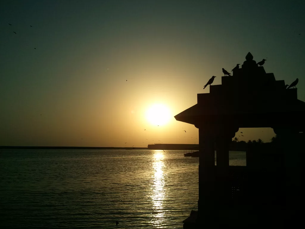 Triveni Sangam near Somnath Temple, Gujarat during sunset, featuring silhouetted temple gate with perched birds, radiant sun reflection on Arabian Sea waters and distant horizon, perfect spiritual landmark with Gujarat tour packages.