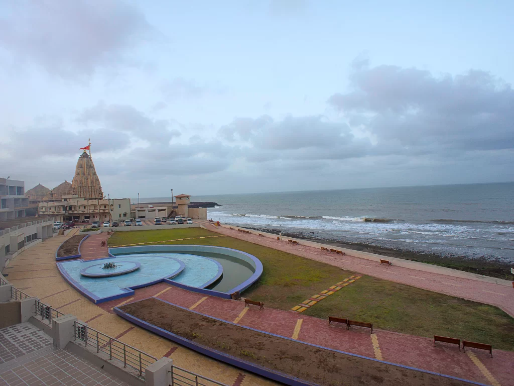 Triveni Sangam plaza near Somnath Temple during overcast monsoon, featuring Somnath Temple with saffron flag, curved blue fountain, landscaped promenade and Arabian Sea horizon, perfect pilgrimage landmark with Gujarat tour packages.