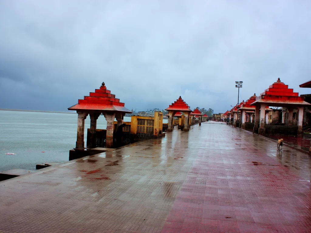 Triveni Sangam Ghat near Somnath Temple during monsoon, featuring red-roofed pavilions along rain-slicked promenade extending to Arabian Sea confluence of Hiran, Kapila and Saraswati rivers, perfect pilgrimage landmark with Gujarat tour packages.