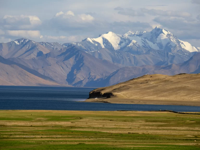 Tso Moriri Lake in Ladakh during partly cloudy day, featuring snow peaks of Zanskar range, turquoise waters and green meadows, perfect adventure experience with Ladakh tour package.