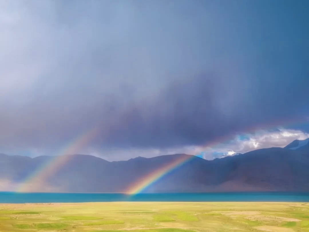 Double rainbow over Tso Moriri Lake in Ladakh during stormy skies, featuring turquoise waters, green meadows and distant mountains, perfect magical adventure with Ladakh tour package. 