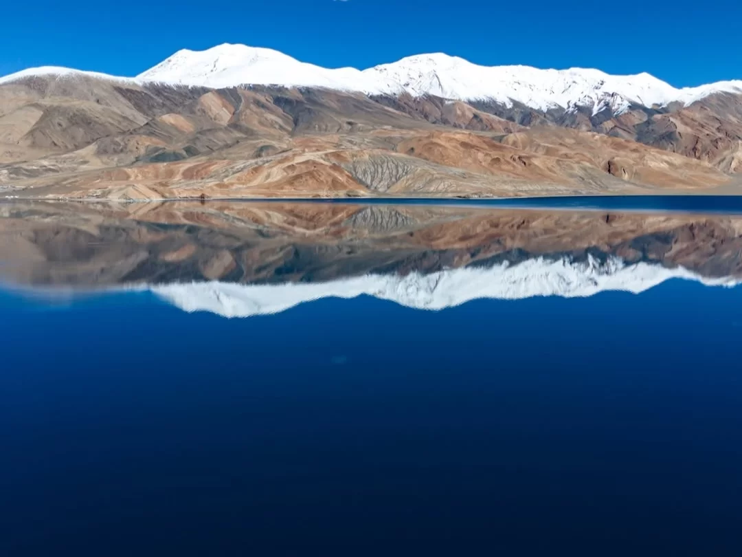 Snow mountains reflecting in Tso Moriri Lake Ladakh during clear blue skies, featuring perfect mirror image on turquoise waters, perfect adventure experience with Ladakh tour package. 