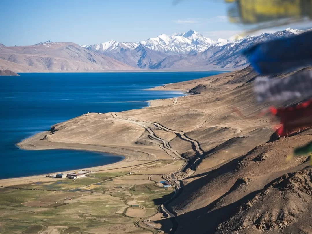Tso Moriri Lake in Ladakh during clear skies, featuring turquoise waters, prayer flags, snow peaks and barren hills, perfect spiritual adventure with Ladakh tour package.