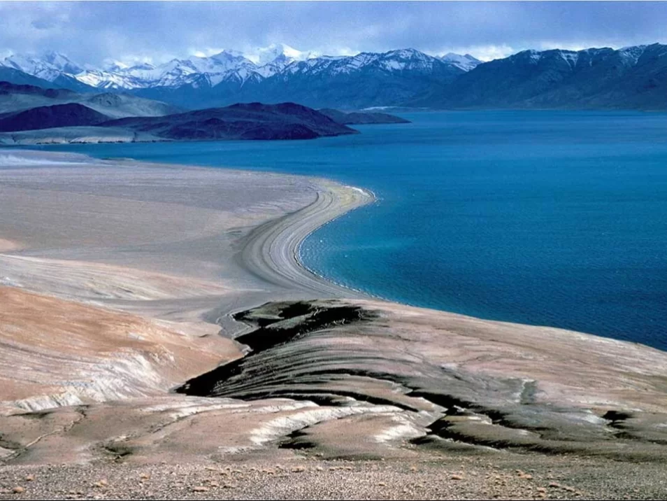 Sandy spit at Tso Moriri Lake in Ladakh during partly cloudy skies, featuring turquoise waters and snow mountains, perfect serene adventure with Ladakh tour package.