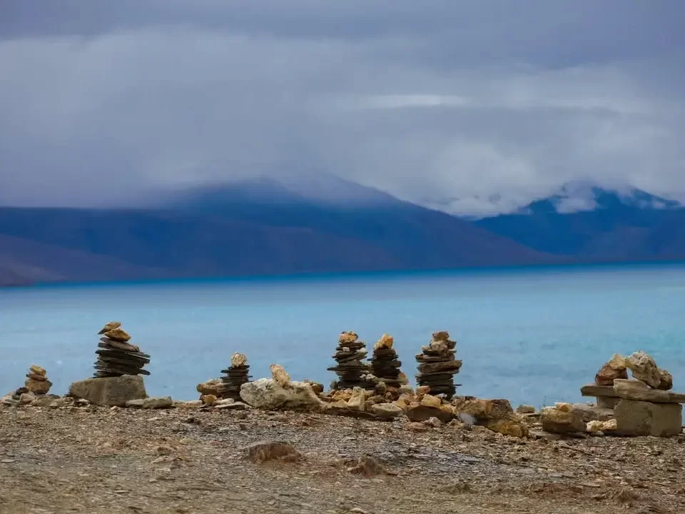 Stone cairns at Tso Moriri Lake in Ladakh during overcast skies, featuring turquoise waters, misty snow mountains and rocky shore, perfect adventure experience with Ladakh tour package. 