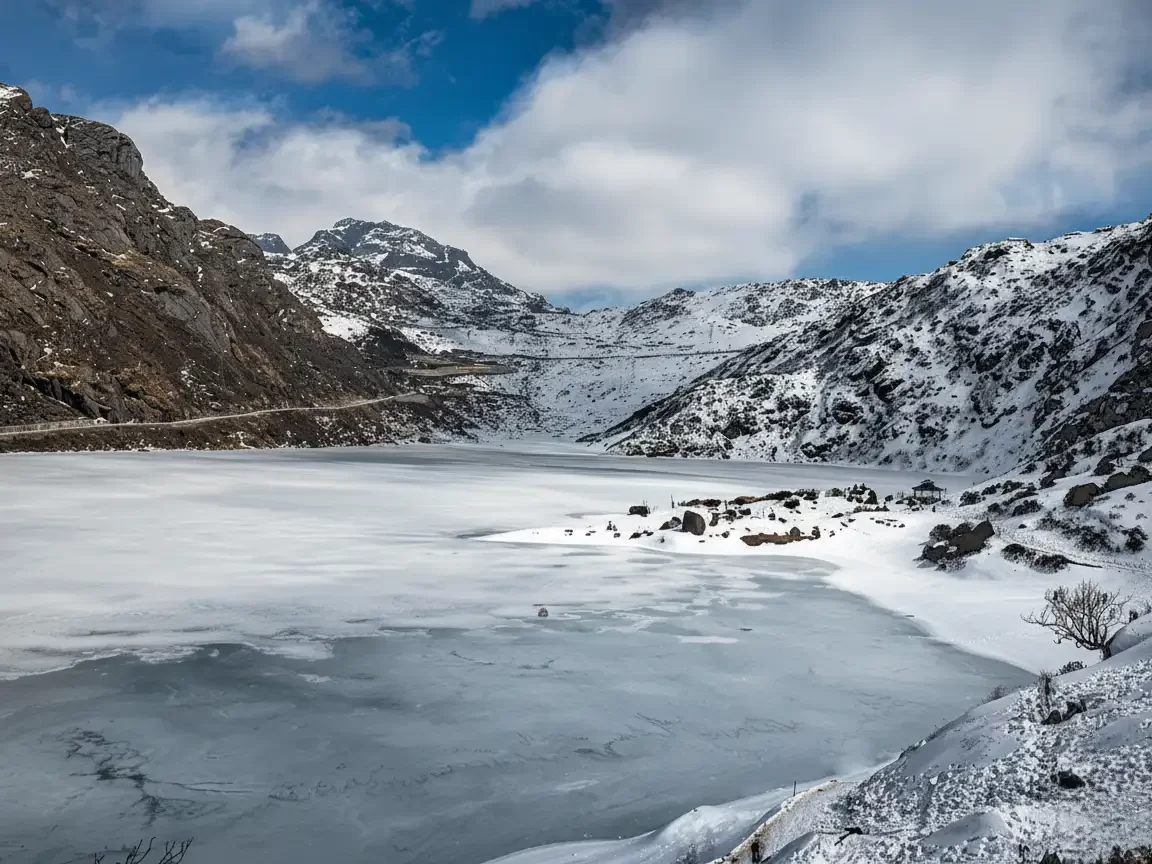 Tsomgo Lake frozen during winter day, featuring icy surface, snowy mountains, Nathula Pass road, blue skies with clouds, perfect adventure Sikkim tour packages.